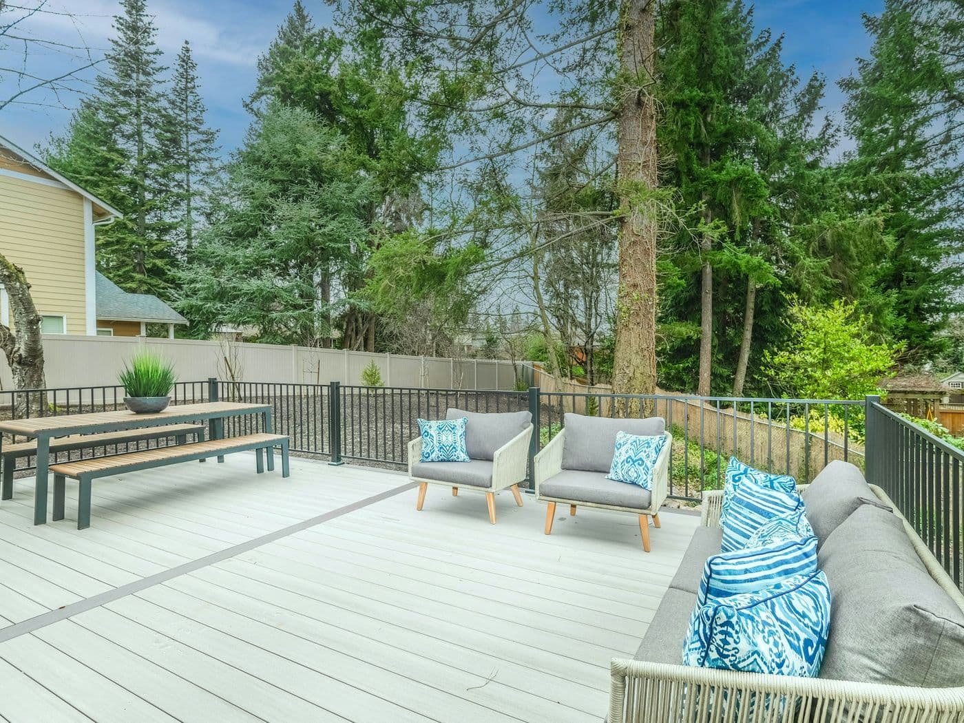 Large pale composite deck with a black metal railing, bench seating and a dining table, backed by conifers