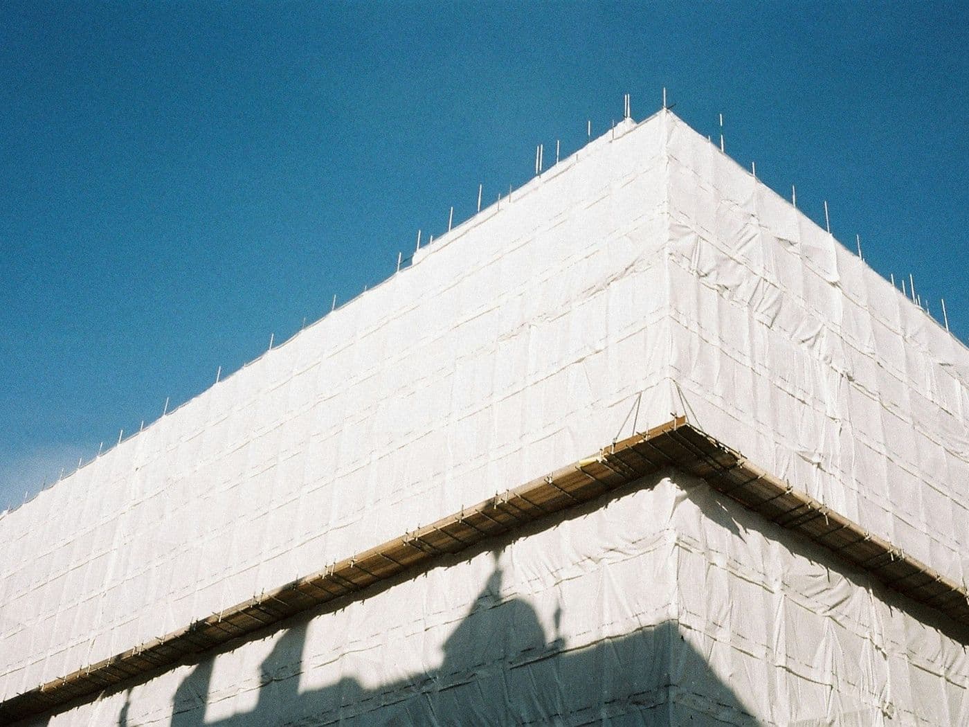 A building corner fully wrapped in white protective sheeting against a clear blue sky, with scaffold decking along the roofline