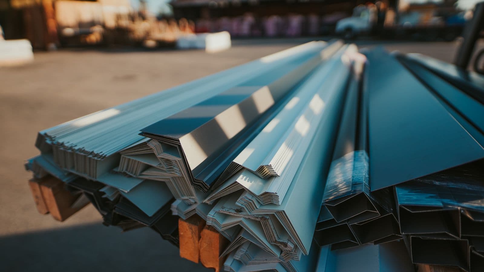 Bundles of custom-bent metal flashing profiles in grey and blue tones stacked on wood blocks in the UBS yard.