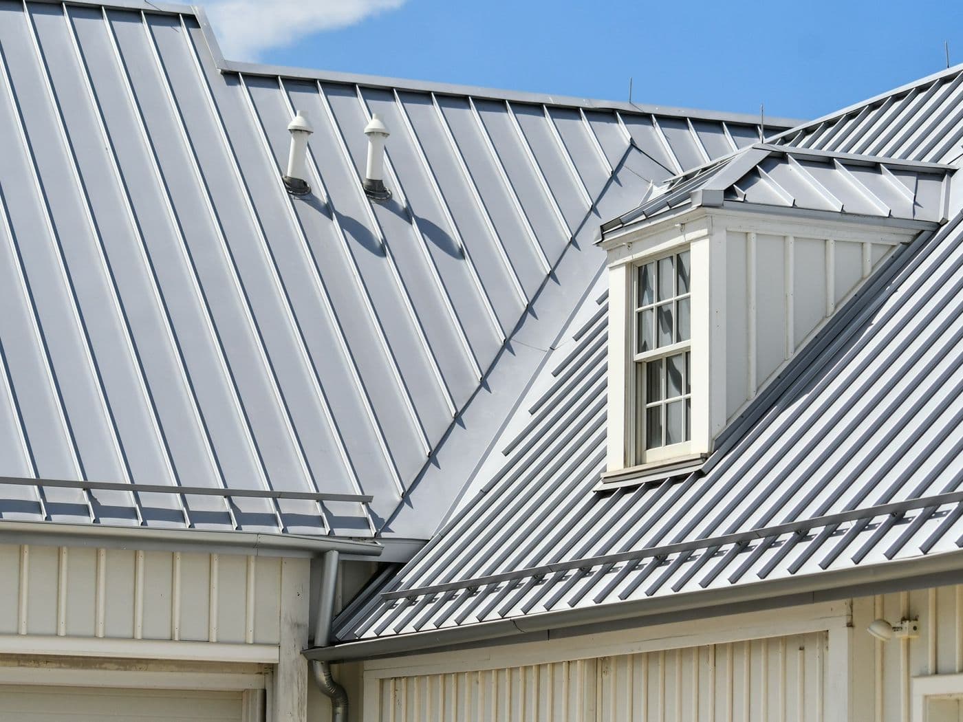 Two grey standing-seam metal roof planes meeting at a valley, with a white dormer and lap-sided wall