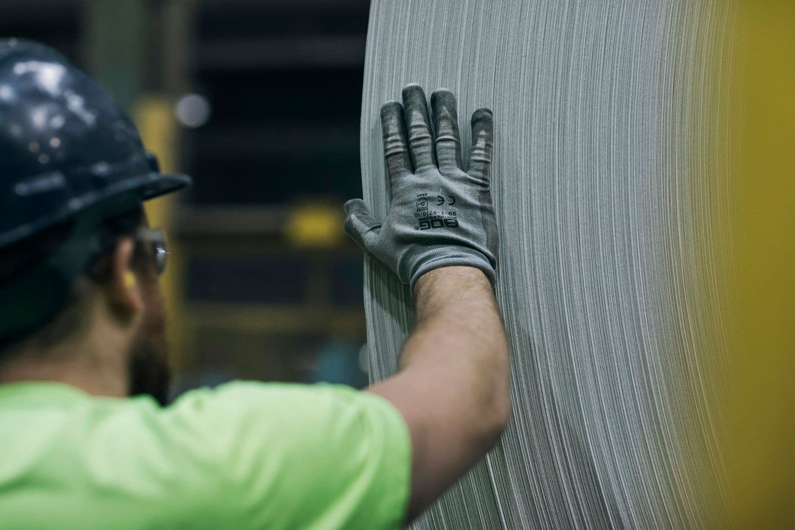 Worker in a hard hat and hi-vis shirt resting a gloved hand against a large coil of sheet metal