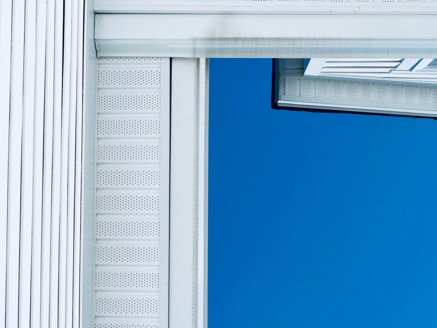 Close-up of a white perforated vented soffit panel and fascia beneath a roof edge, against a deep blue sky