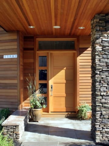 Home entryway with a cedar tongue-and-groove soffit overhead and cedar channel siding beside a wood front door.
