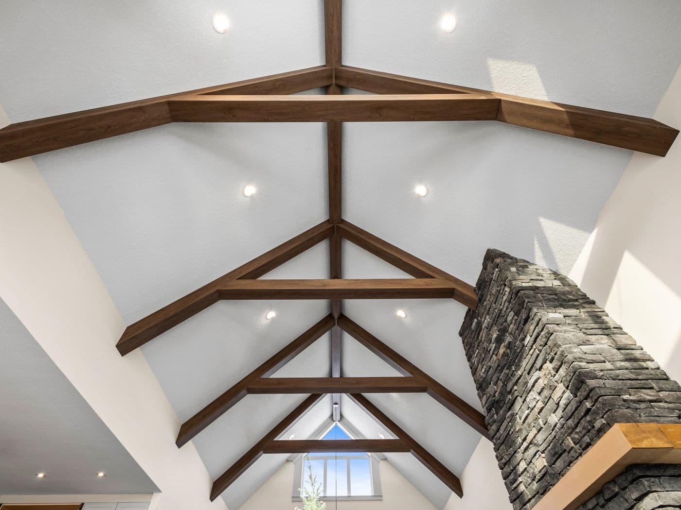 Vaulted interior ceiling with dark wood-tone faux ceiling beams and trusses against white walls, beside a stacked stone chimney.