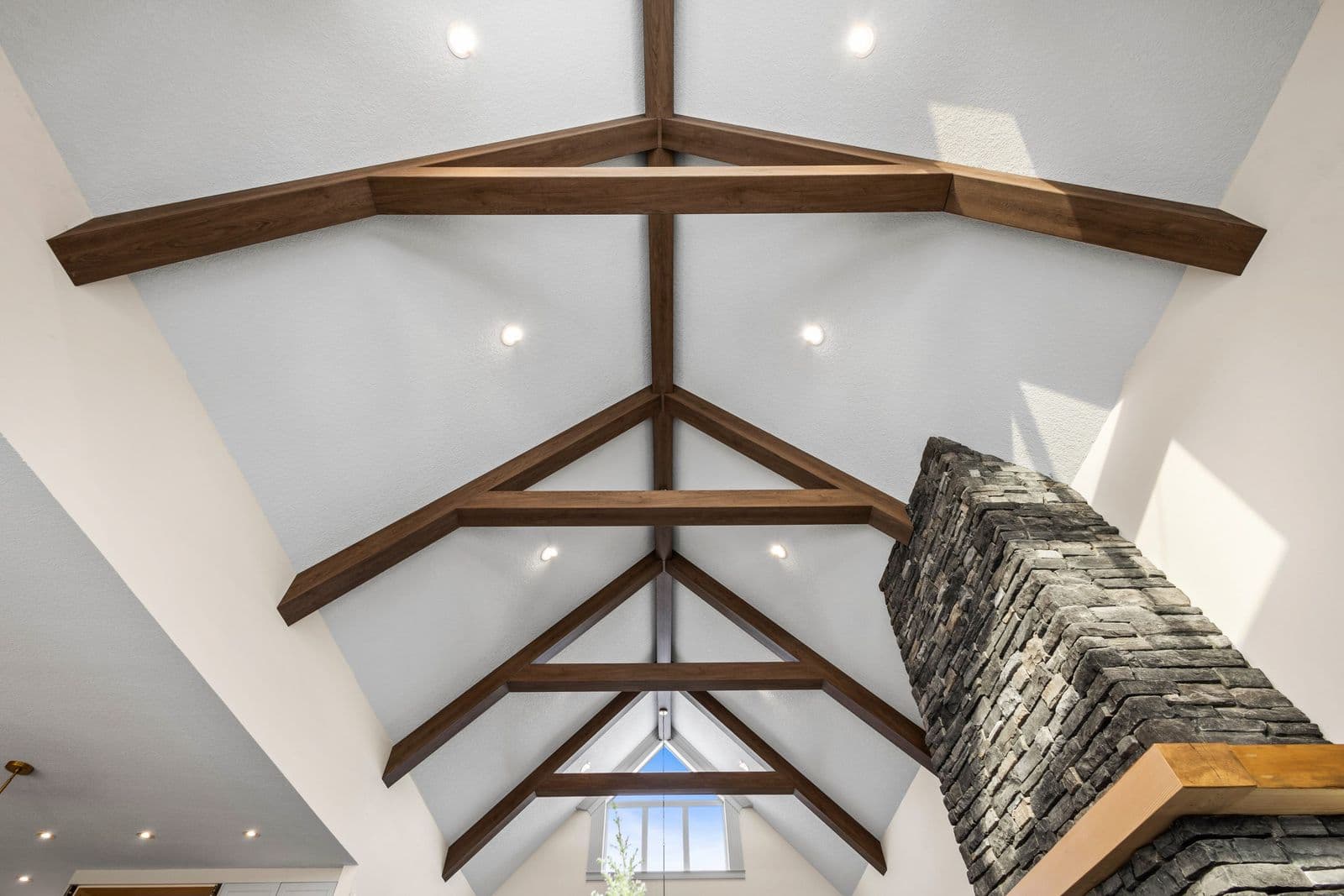 Vaulted interior ceiling with dark wood-tone faux ceiling beams and trusses against white walls, beside a stacked stone chimney.