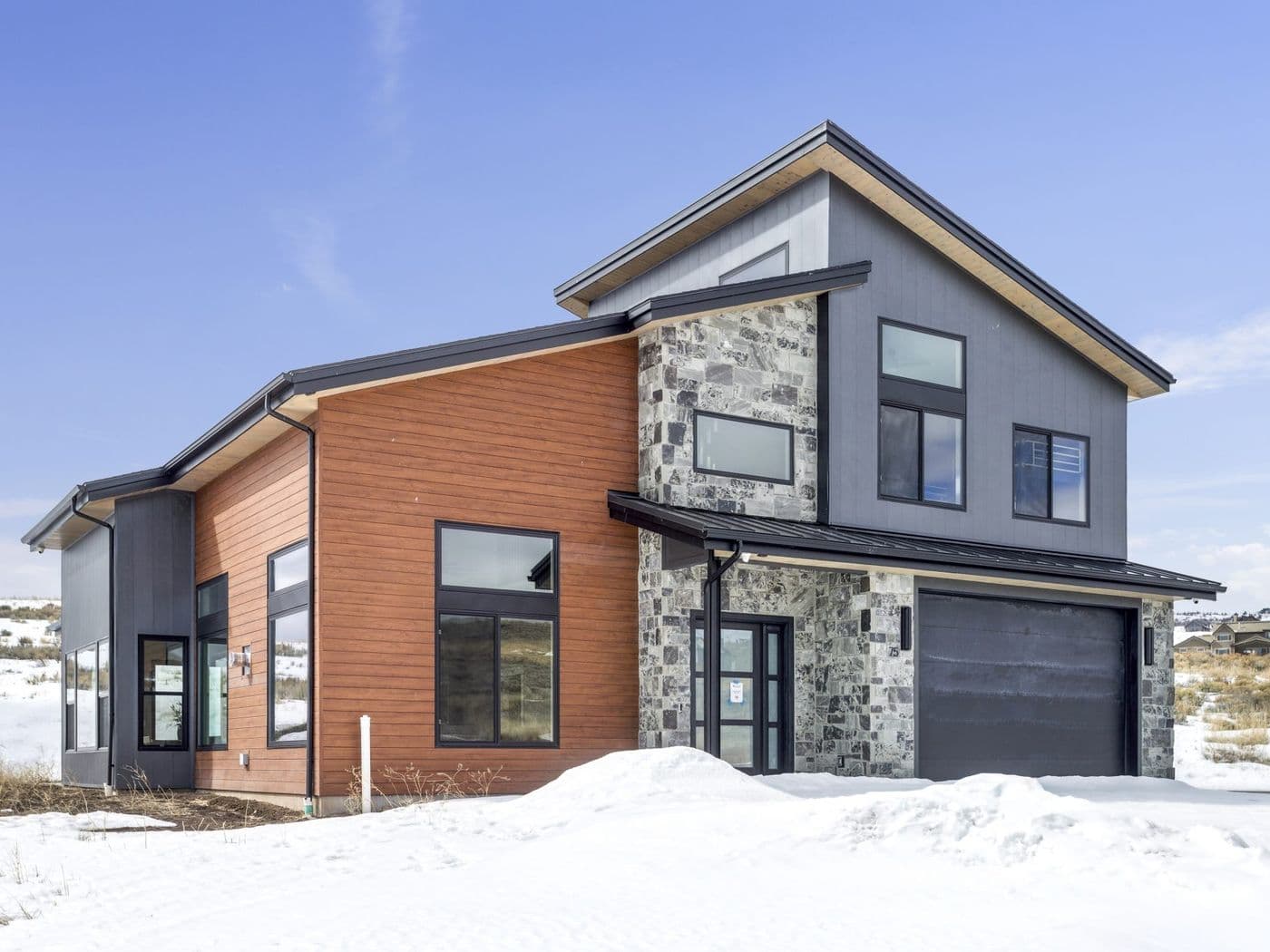 Modern two-storey home in snow with reddish-brown horizontal wood-look wall panel siding, grey stone veneer accents, and black-framed windows.
