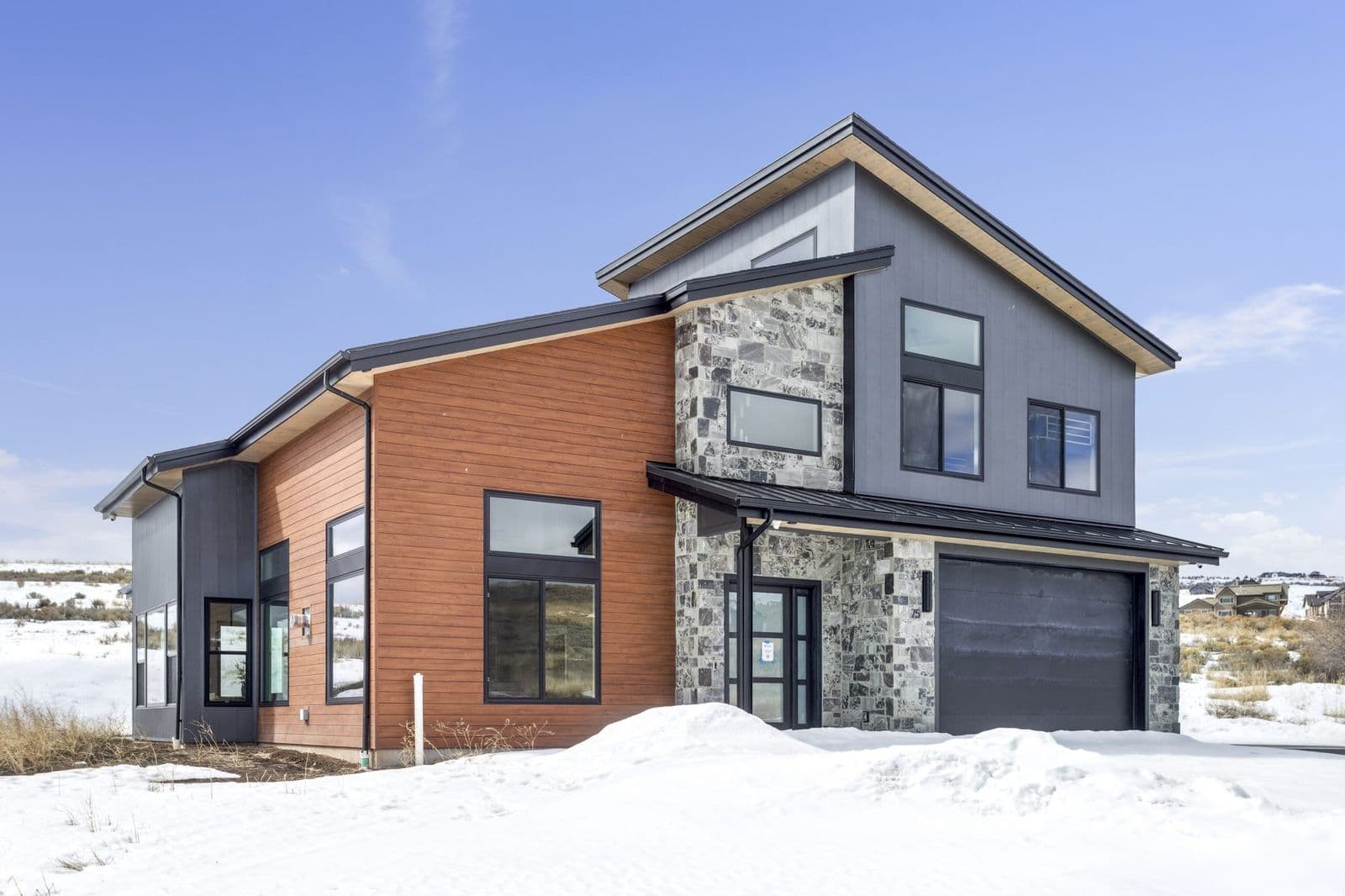 Modern two-storey home in snow with reddish-brown horizontal wood-look wall panel siding, grey stone veneer accents, and black-framed windows.