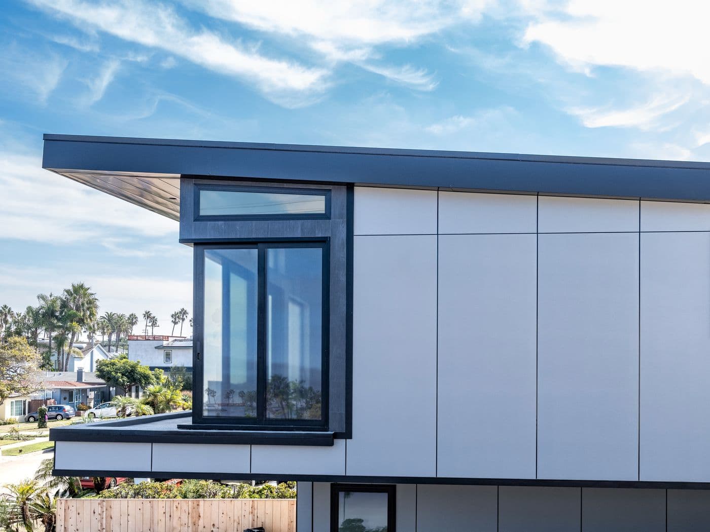 Close-up of a modern home's roofline and window corner, clad in white vertical flush-panel siding with visible seams, palm trees in the background.