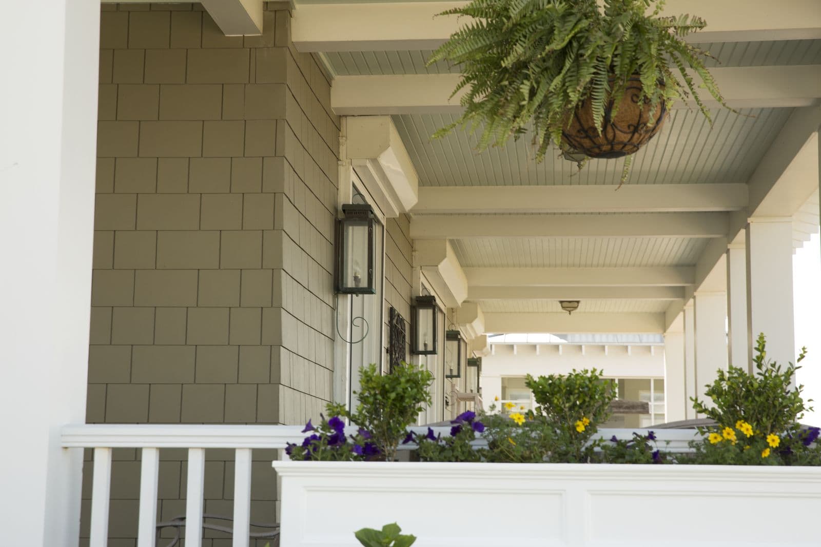 Craftsman porch with sage-green coursed shingle siding on the wall and column pier, a hanging fern basket, and wall-mounted lanterns.