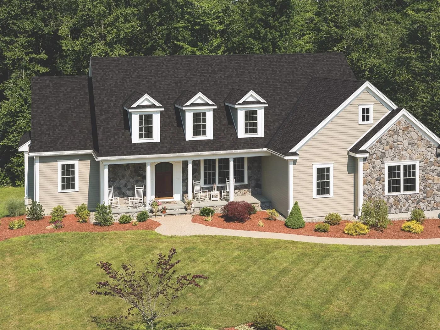 Aerial view of a beige single-storey house with a dark Dual Black IKO Cambridge shingle roof, three front dormers, and a stone-accented gable.