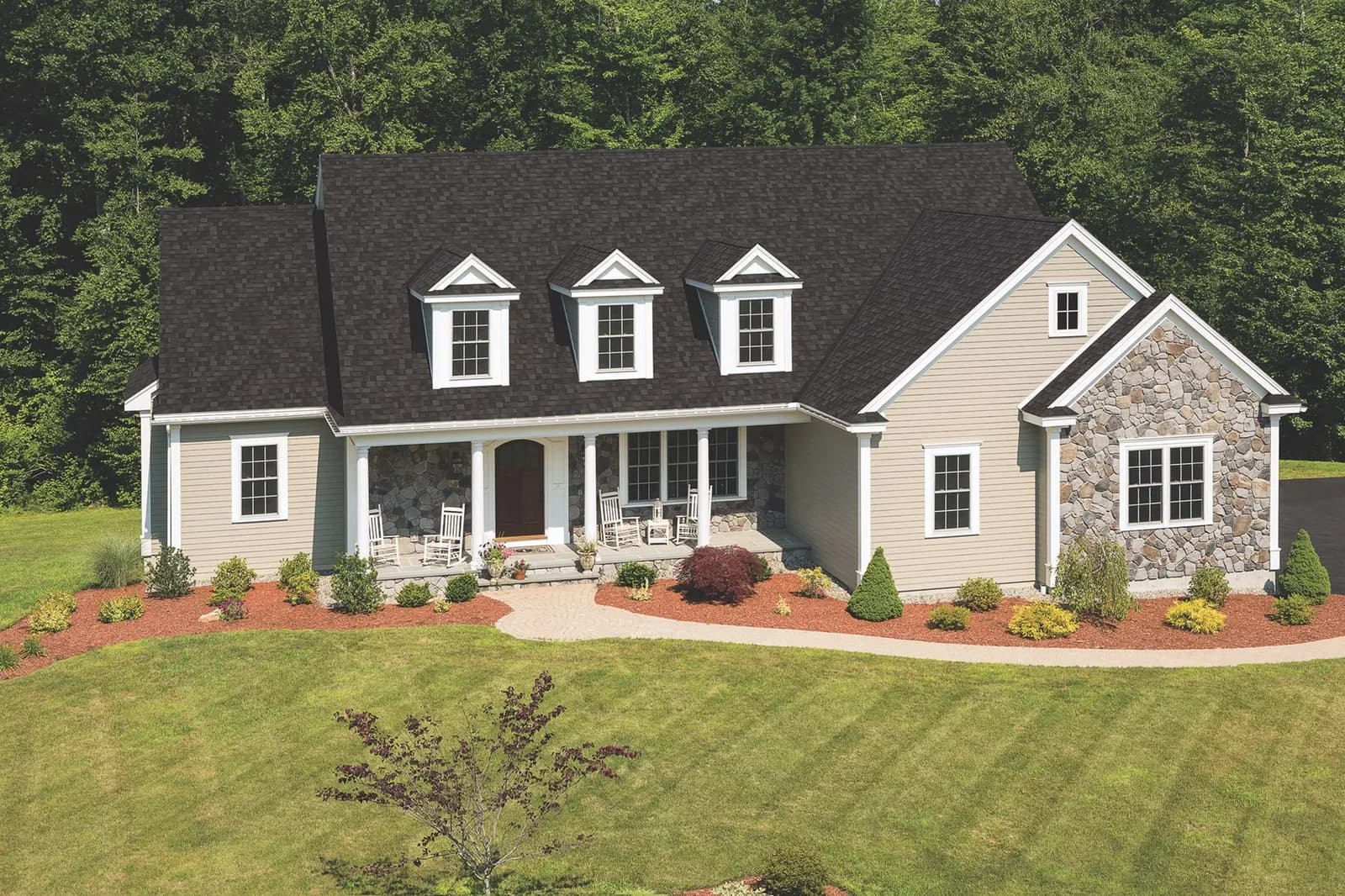 Aerial view of a beige single-storey house with a dark Dual Black IKO Cambridge shingle roof, three front dormers, and a stone-accented gable.