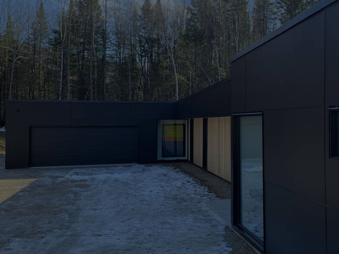 Dusk photo of a modern home with black vertical panel siding and a light wood-toned window surround, snow on the ground.