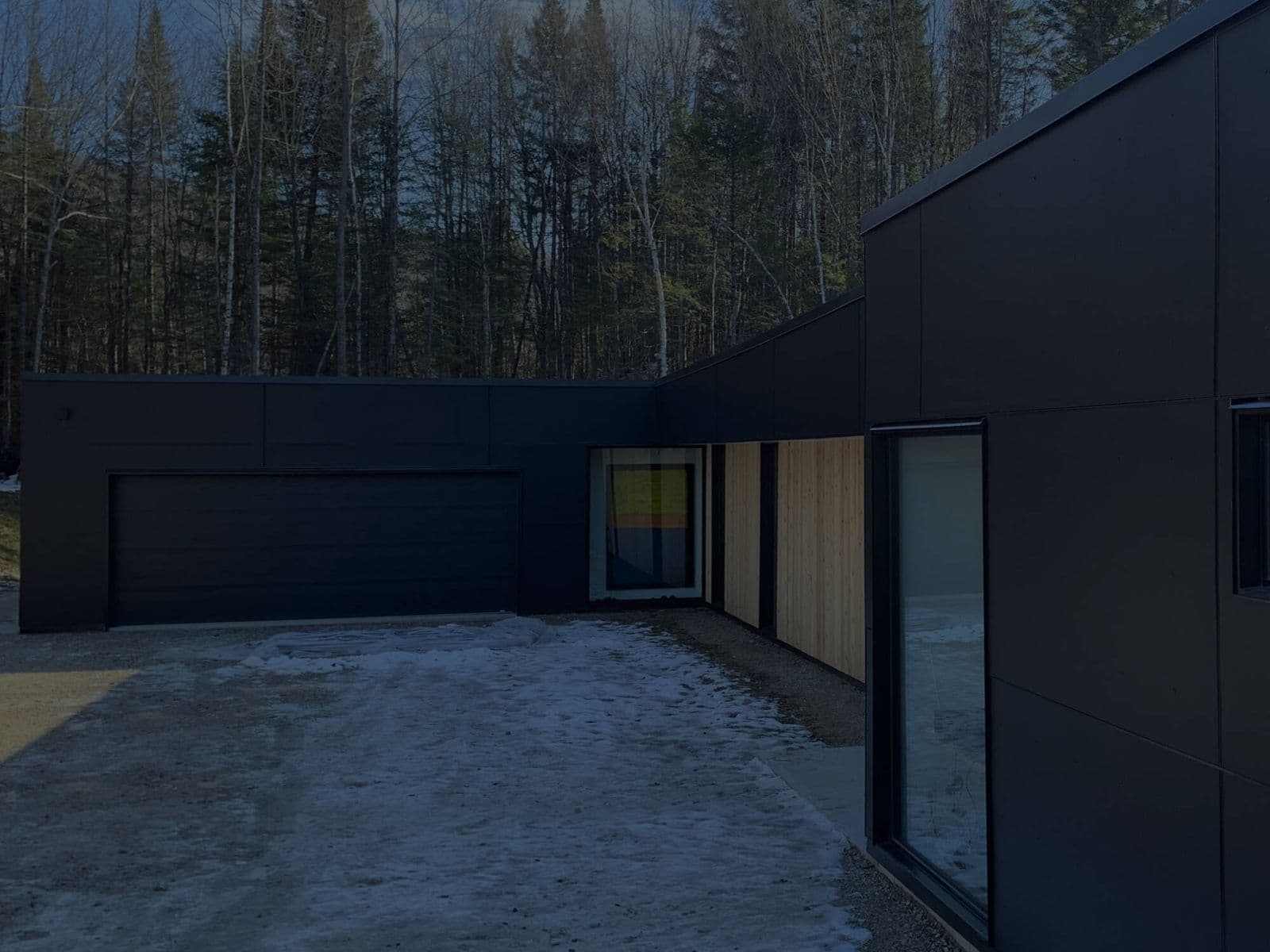Dusk photo of a modern home with black vertical panel siding and a light wood-toned window surround, snow on the ground.