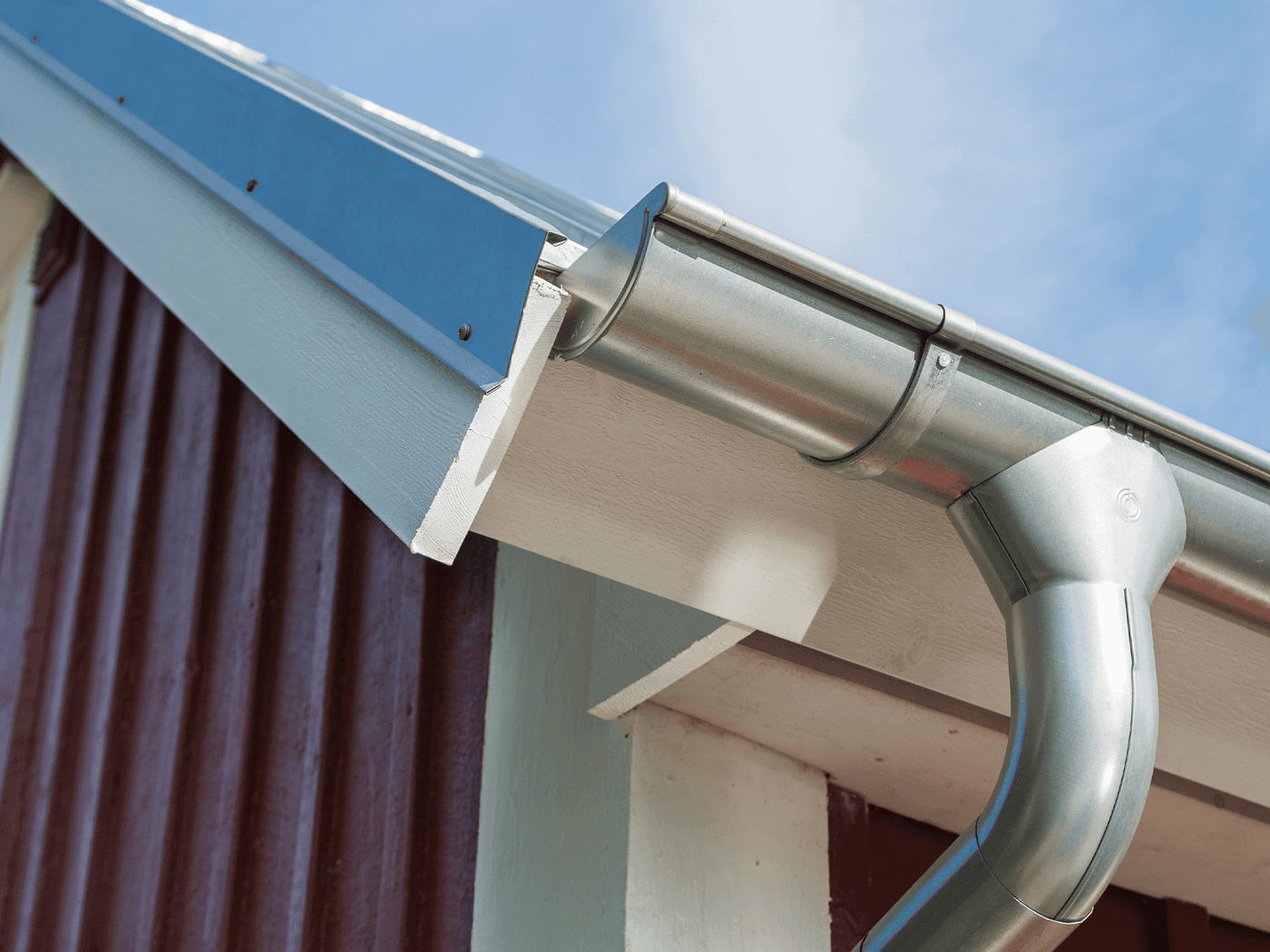 Close-up of a galvanized steel half-round gutter at a roofline corner, with a downpipe elbow curving down past white fascia and dark wood siding.