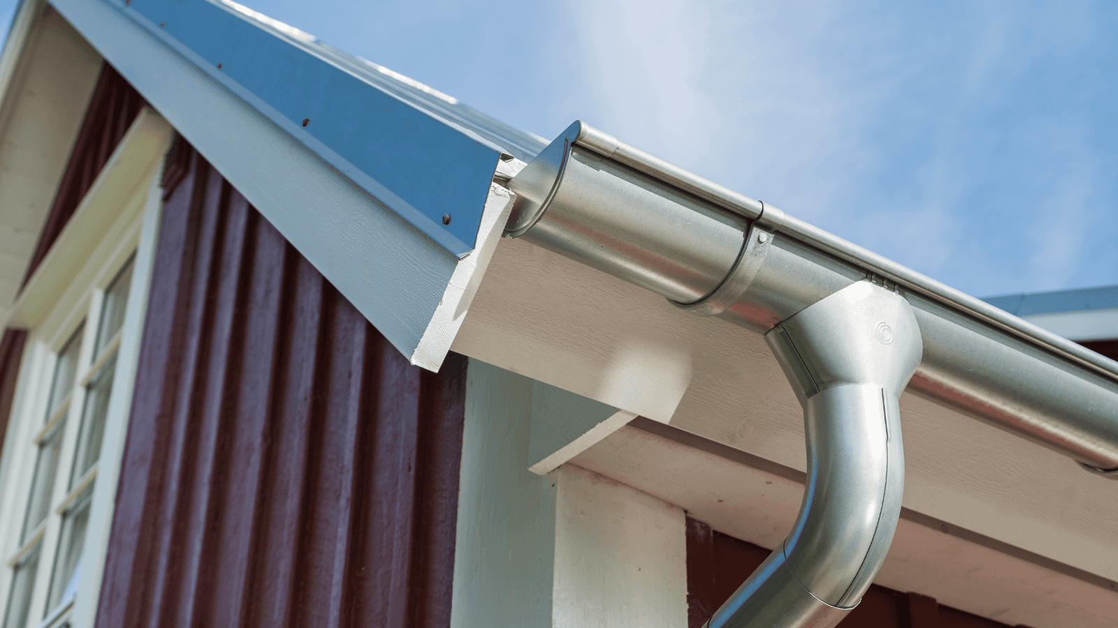 Close-up of a galvanized steel half-round gutter at a roofline corner, with a downpipe elbow curving down past white fascia and dark wood siding.