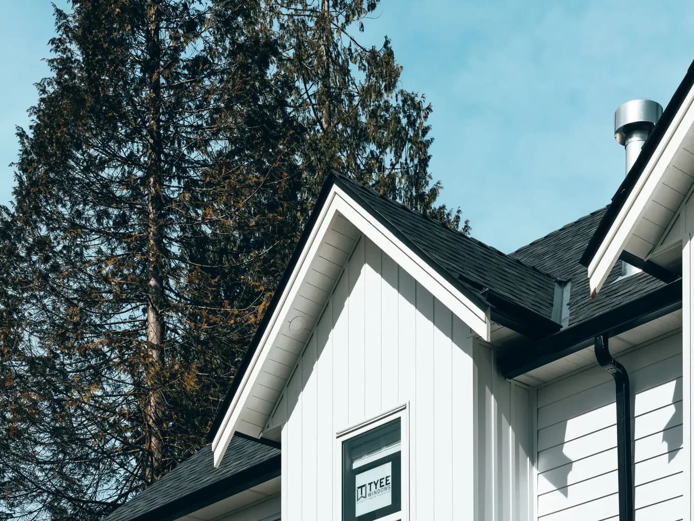 Gable end of a home with white vertical board-and-batten and horizontal lap aluminum siding, black roofline, against evergreen trees and a blue sky.