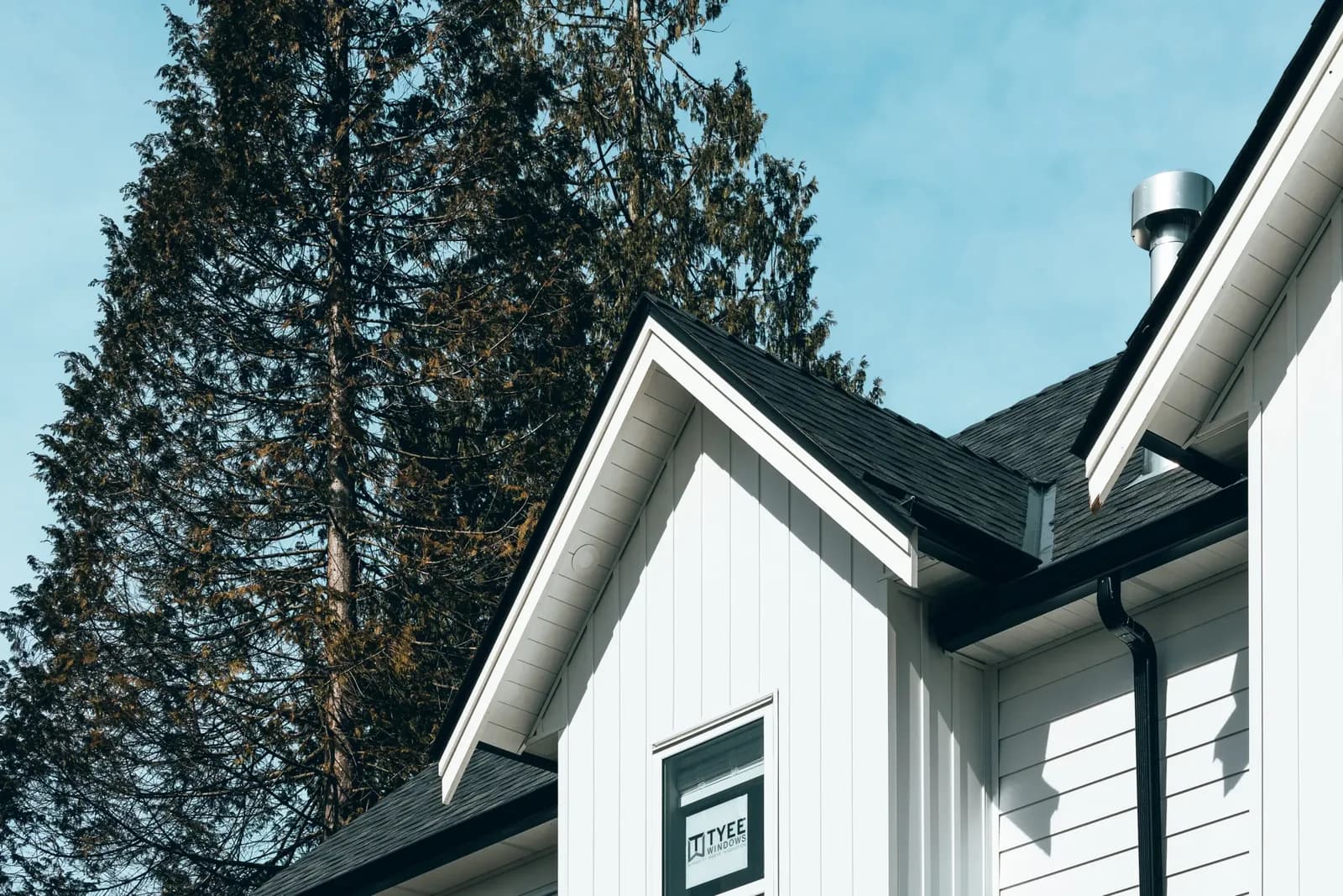 Gable end of a home with white vertical board-and-batten and horizontal lap aluminum siding, black roofline, against evergreen trees and a blue sky.
