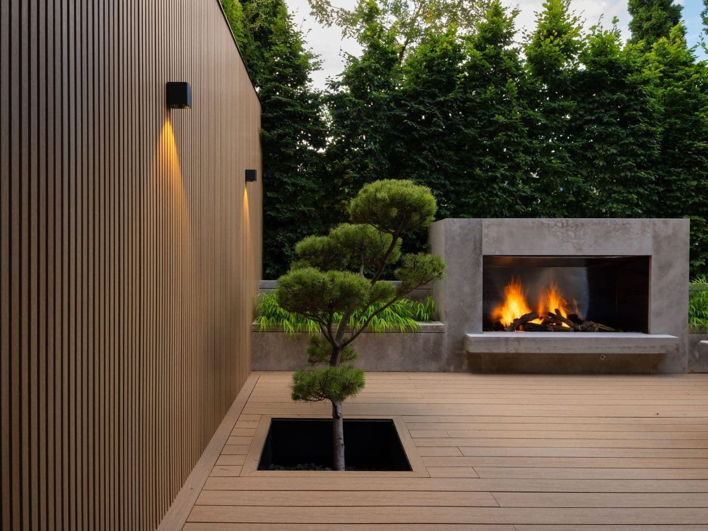 Dusk photo of a modern building's vertical wood-tone fluted composite siding, a built-in concrete outdoor fireplace, and a matching composite deck.