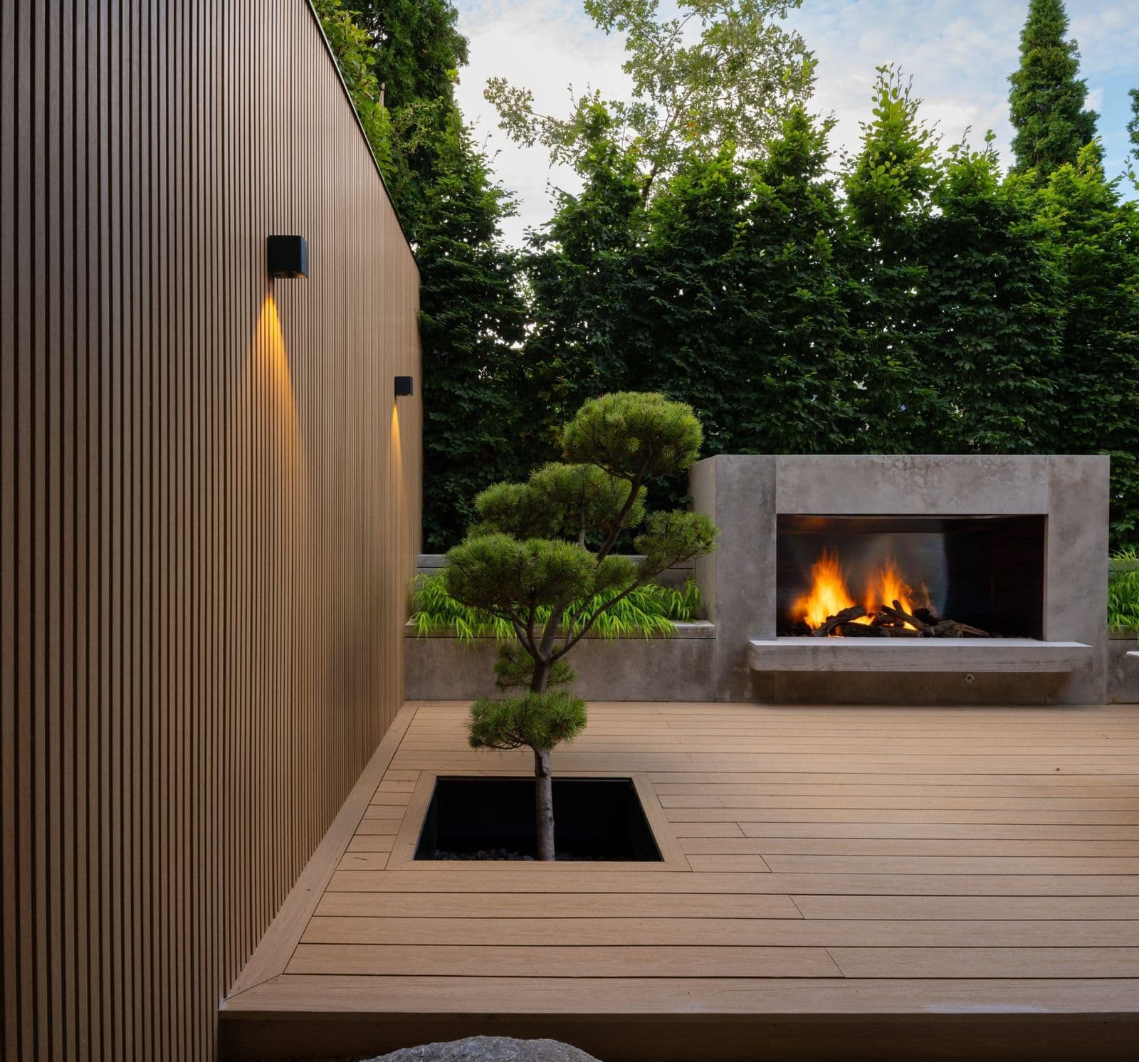 Dusk photo of a modern building's vertical wood-tone fluted composite siding, a built-in concrete outdoor fireplace, and a matching composite deck.