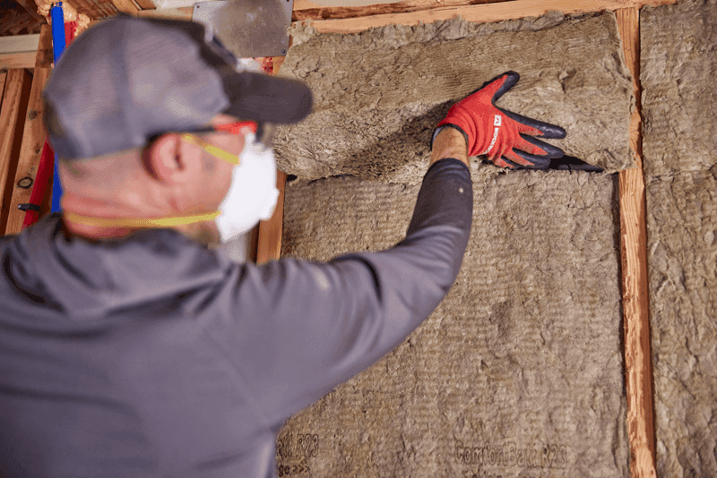 Installer in gloves and a dust mask press-fitting ComfortBatt stone wool insulation between wood wall studs.