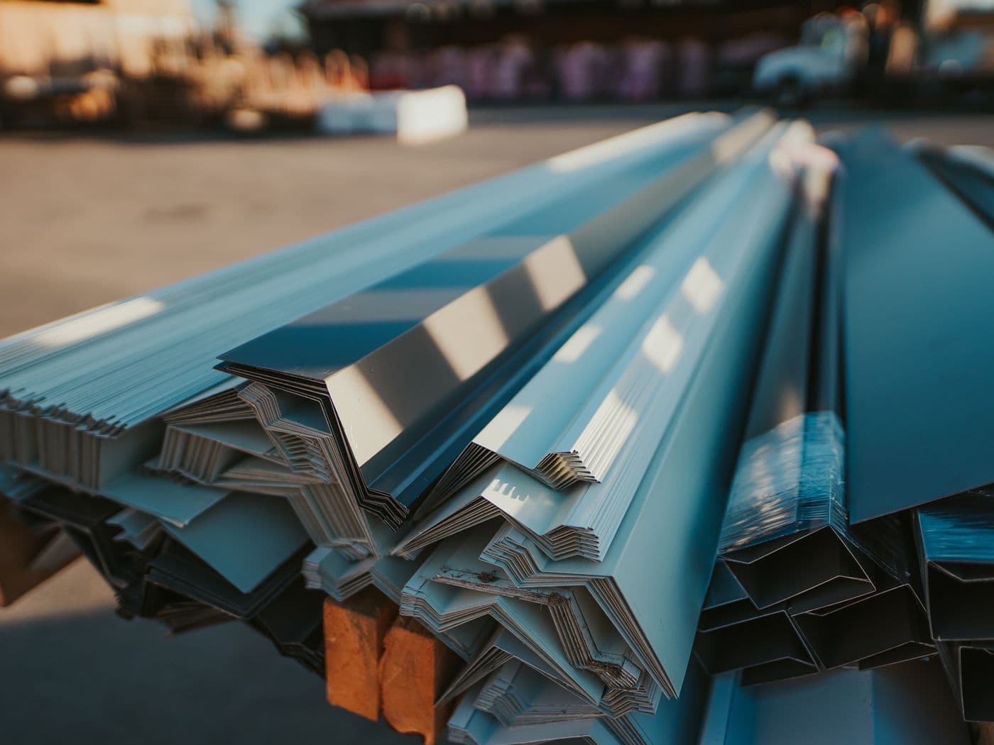 Bundles of custom-bent metal flashing profiles in grey and blue tones stacked on wood blocks in the UBS yard.