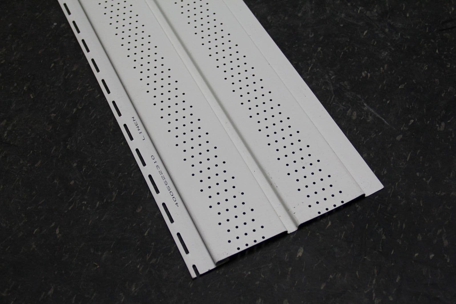 Close-up of a single white vented vinyl soffit panel on a dark stone surface, showing rows of perforated vent holes and a stamped "Linen" colour name near the nailing hem.